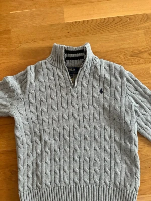 Grå kabelstickad tröja Polo Ralph Lauren - Snygg grå kabelstickad tröja från Polo Ralph Lauren med halv dragkedja och hög krage. Tröjan har ribbade muddar och det klassiska broderade logotypen på bröstet. Perfekt för lager på lager och en stilren look. Passar dig som är mellan 145/155cm
