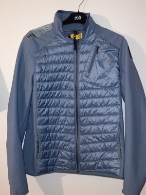 Blå vindjacka från Parajumpers - Snygg blå vindjacka från Parajumpers med quiltad front, dragkedja och bröstficka. Jackan har ett coolt patch på ärmen och hög krage med gul detalj vid halsen. Perfekt för dig som vill ha en lätt och stilren jacka till vardags.