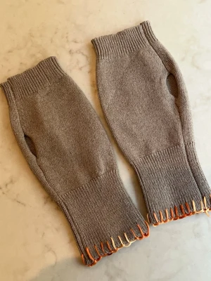 handledsvärmare cashmere - Säljer ett par beiga stickade handledsvärmare med tumhål och ribbade muddar. De har handsydda detaljer i orange och gult. Perfekta för att hålla händerna varma och ändå kunna använda mobilen. Otroligt mjuka i 100% cashmere.