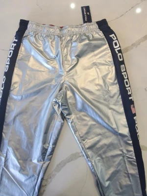 Silver mjukisbyxor från Polo Sport - Snygga silverfärgade mjukisbyxor från Polo Sport med marinblå sidor och vit logga längs benen. Byxorna har resår i midjan och dragsko för skön passform. Perfekta för dig som vill sticka ut med en sportig och futuristisk look.