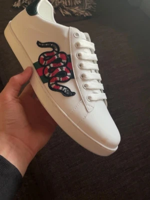 Gucci Ace skor - (TAR ÄVEN BYTE MOT MAISON MARGIELA)Säljer ett par vita Gucci Ace sneakers i läder med den ikoniska broderade ormen på sidan och gröna/röda detaljer. Klassisk låg modell med vita snören och präglad sula med Gucci-logga undertill. Perfekta för dig som vill sticka ut med lyxig streetstyle. 