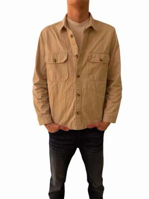 Beige overshirt med bröstfickor - Stilren beige overshirt med klassisk krage och två stora bröstfickor med knappar. Tillverkad i ett mjukt bomullstyg och har knäppning framtill. Perfekt att bära över en t-shirt för en avslappnad look.