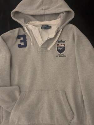 Grå Polo Ralph Lauren Hoodie - Snygg grå hoodie från Polo Ralph Lauren med broderad logga och texten 'POLO RL 867 MADISON' på bröstet. Har en stor huva med dragsko, känguruficka och mörkblå siffra 3 på axeln. Mjuk bomullsmix och vita detaljer vid knappar och snören.