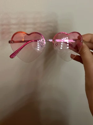 Hjärtformade rosa solglasögon - Säljer ett par riktigt söta solglasögon med hjärtformade glas i transparent rosa plast. Bågarna är också rosa och ger en lekfull vibe till din outfit. Perfekta för dig som vill sticka ut och addera lite färg till sommaren.