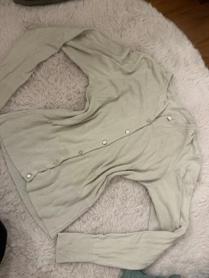 Ljusbeige ribbad kofta med knappar - Ljusbeige kofta med långa ärmar och ribbade detaljer. Koftan har en rak passform och knäpps framtill med små vita knappar. Perfekt att slänga över en t-shirt eller linne för en chill look. Obs e lite smutsig på ärmen