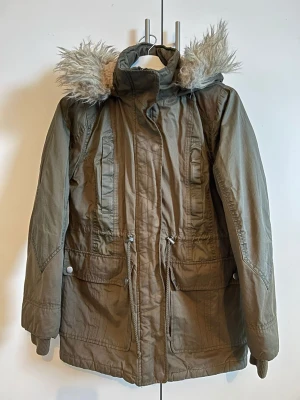 TU Parkas i Khaki 🌿✨ - Superfin khakigrön parkas från TU! Perfekt för kalla dagar då den är både varm och praktisk. ❄️ • Storlek: 40 (UK 12) • Detaljer: Mysig fuskpäls på huvan, stora fickor och dragsko i midjan så man kan justera passformen. 