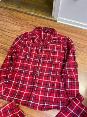 Röd rutiga pyjamas i flanell - Mysig pyjamas i röd flanell med klassiskt rutigt mönster i svart och vitt. Setet består av en långärmad skjorta med knappar och bröstficka samt matchande byxor med resår i midjan. Perfekt för chilliga kvällar hemma.