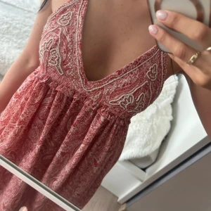 Röd paisleymönstrad klänning från Zara - Supersöt röd och vit kortklänning från Zara med paisleymönster och djup v-ringning. Klänningen är ärmlös och har en luftig passform, perfekt för varma dagar. Tillverkad i mjuk bomull och har en bohemisk vibe som passar perfekt till sommaren.