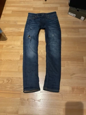 Blå slitna Jack and Jones jeans med raka ben - Säljer ett par blå jeans med raka ben och slitningar på låren och knäna. Jeansen har klassisk femficksdesign, normal midja och snygga slitdetaljer framtill. Materialet är denim i bomull och de har en läderpatch bak i midjan.