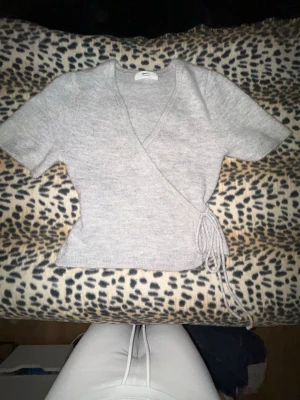 Grå omlottopp med knytband - Snygg grå omlottopp med kort ärm och knytband i sidan. Mjuk stickad känsla och djup v-ringning.aldrig blivit använd efter köpet