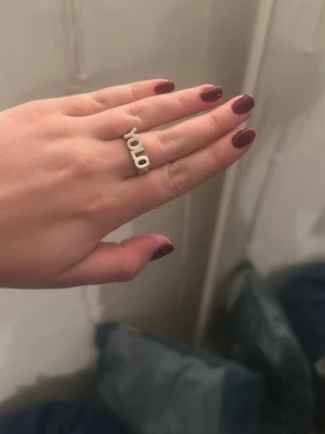 Silvrig YOLO-ring i modern design från L8h - Säljer en cool ring i silverfärgad metall med texten 'YOLO' i stora bokstäver. Ringen har en öppen, modern design och passar perfekt för dig som vill sticka ut med ett statement-smycke.
