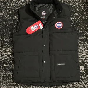Svart dunväst från Canada Goose - Snygg svart dunväst från Canada Goose med klassisk logga på bröstet och två stora fickor framtill. Västen har hög krage och knäppning med tryckknappar. Perfekt för kalla dagar när du vill hålla stilen.