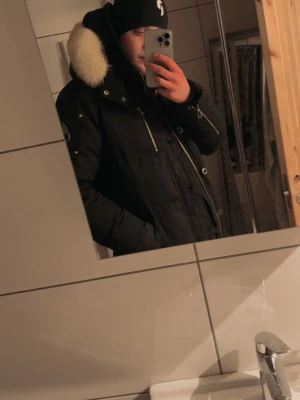 Moose 3q  - Snygg svart parkasjacka med stor huva och fluffig ljus pälskrage. Jackan har dragkedja framtill, flera fickor och en klassisk parkasdesign. Perfekt för kyliga dagar och ger en stilren look.