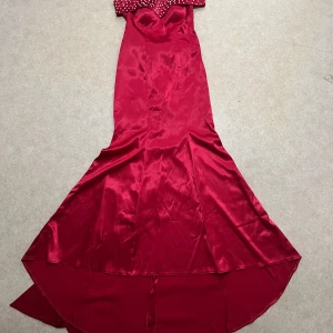 Röd långklänning med rosett - Elegant röd axelbandslös långklänning i satin med en stor dekorativ rosett och glittriga detaljer. Perfekt för en glamorös kväll.  Jag säljer den på grund av ändrad storlek. Klänningen är perfekt för bal, bröllop och olika sorters fester. 