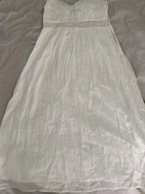 Vit långklänning med spetsdetaljer - Romantisk vit långklänning med tunna axelband och v-ringad hals. Klänningen har fina spetsdetaljer och broderade blommor upptill samt ett skirt, luftigt tyg i kjolen. Perfekt för sommarens alla tillfällen.