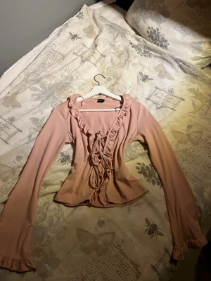 Ljusrosa blus med volang och snörning - Supersöt ljusrosa blus med långa ärmar, volangdetaljer längs ringningen och snörning framtill. Figurnära passform och lätt utsvängda ärmslut. Från Gina i str S