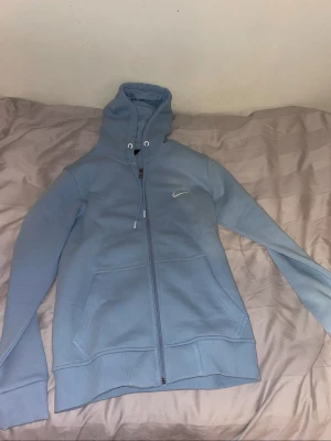 Blå och röd Nike hoodie med dragkedja - Två snygga hoodies från Nike, en i ljusblått och en i rött med svarta detaljer. Båda har dragkedja framtill, huva med dragsko och fickor. Den röda har extra ficka på ärmen och coola svarta linjer. Perfekt för dig som vill ha sportig och stilren look.