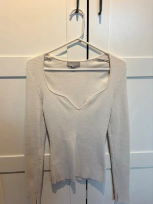 Beige ribbstickad tröja med v-ringning - Snygg beige ribbstickad tröja med unik v-ringad hals och långa ärmar. Tröjan har en liten slits vid ärmslutet och är tillverkad i ett mjukt, stretchigt material. Perfekt för lager på lager och enkel att matcha med olika outfits.