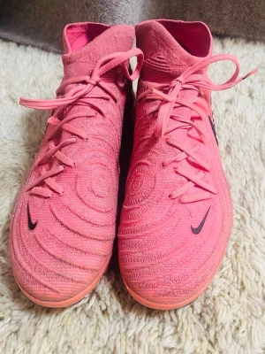 Rosa Nike fotbollsskor med snörning - Säljer ett par snygga rosa Nike fotbollsskor med hög strumpa och snörning. Sulan är rosa med dobbar för bra grepp på planen. Perfekta för dig som vill sticka ut på fotbollsplanen!