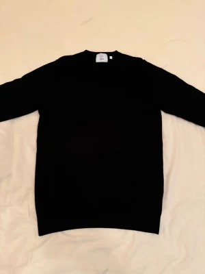Bläck Tröja - En stilren svart tröja i bomull från Bläck med rund halsringning och långa ärmar. Tröjan har en klassisk passform och är perfekt för dig som gillar enkel och tidlös design. Materialet ser mjukt och bekvämt ut, perfekt för lager på lager.