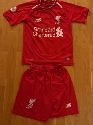 Liverpool matchställ M. Salah 11 - Röd Liverpool matchställ med tröja och shorts från New Balance. Tröjan har tryck med Standard Chartered, klubbmärke och M. Salah 11 på ryggen. Shorts med LFC-logga och New Balance-märke. Perfekt för fotbollsträning eller supporterliv.