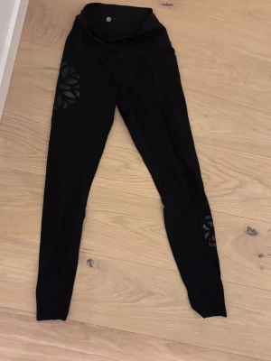 Svarta leggings med blommönster - Snygga svarta leggings med hög midja och ett stilrent blommönster i blankt material på ena benet. Perfekta för träning eller chill, med stretchigt tyg som sitter skönt. Passar dig som gillar enkel men ändå unik stil.