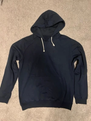 Mörkblå hoodie från Pier one  - En stilren mörkblå hoodie med huva och vita dragsnören. Hoodien har en klassisk känguruficka framtill och ribbade muddar vid ärmslut och nederkant. Perfekt för en avslappnad och trendig look.