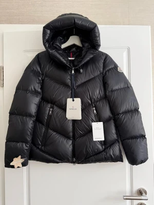 Svart Moncler dunjacka med huva - Snygg svart dunjacka från Moncler med huva och dragkedja framtill. Jackan har två snedställda fickor med dragkedja och en dekorativ stjärndetalj på ena ärmen. Perfekt för kalla dagar och riktigt trendig look.