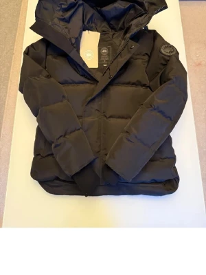 Svart pufferjacka med pälsluva - Snygg svart pufferjacka från Canada Goose med stor huva och fluffig pälsdetalj. Jackan har quiltad design, dragkedja och knappar framtill samt ribbade muddar vid ärmslut. Perfekt för kalla vinterdagar och har en cool badge på ärmen.Säljer på grund av köpt fel storlek.