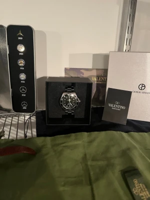 Grisch klocka med svart urtavla - Stilren klocka från R@lex med svart urtavla och silverfärgad länk i metall. Klockan har en klassisk rund form, tydliga siffror på vridringen och datumvisning. Perfekt accessoar för dig som gillar exklusiv och tidlös design.