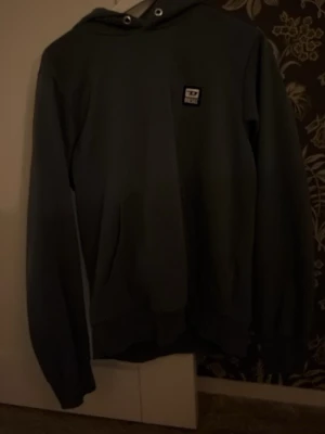 Mörk grön hoodie från diesel - Mörkgrön hoodie med huva och , samt en liten fyrkantig logga på bröstet. Klassisk känguruficka framtill och ribbade muddar vid ärmar och nederkant. Perfekt för en avslappnad och stilren look.