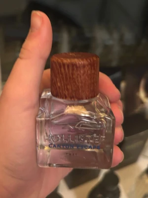 Hollister Canyon Escape parfym - Fräsch parfym från Hollister, Canyon Escape, i en genomskinlig glasflaska med träfärgat lock. Flaskan har en stilren och modern design med blå text och en ingraverad logga på framsidan. Perfekt för dig som gillar en clean och ungdomlig doft. Använd 1 gång! 