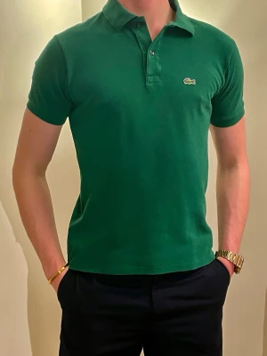 Grön pikétröja från Lacoste - Klassisk grön pikétröja från Lacoste med korta ärmar och den ikoniska krokodilloggan på bröstet. Storlek 164cm. Tröjan har krage och knappar vid halsen. Tillverkad i bomull för en bekväm och snygg look.