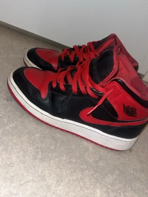 Nike Air Jordan 1 svart/röd sneakers - Säljer ett par klassiska Nike Air Jordan 1 sneakers i svart och rött skinn med röda snören och vit sula. Ikonisk high-top modell med rund tå och snygga detaljer som Jumpman-logga på plösen och Air Jordan-vingen på sidan. Perfekta för dig som gillar streetwear.