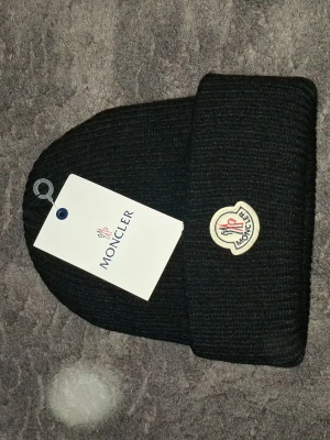 Svart Moncler mössa med logga - Snygg svart ribbstickad mössa från Moncler med uppvikt kant och klassisk Moncler-logga framtill. Perfekt för dig som vill ha en stilren accessoar med exklusiv känsla. Mössan är i mjukt material och passar till de flesta outfits. OBS! Helt ny bara utplockad från påsen.