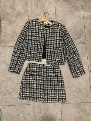 Svartvit rutig kostym med kjol - Snygg svartvit rutig kostym med croppad kavaj och matchande kjol. Kavajen har långa ärmar och raka axlar, kjolen har dekorativa knappar framtill. Klassisk tweed-look som passar perfekt till trendiga outfits.