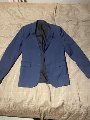 Snygg kavaj från J. Lindeberg - Snygg blå kavaj med två knappar framtill och klassiska fickor med lock. Kavajen har en stilren passform och helfodrad insida.  Den är knappt använd då användaren växte ur den. För fler frågor och funderingar är det bara att kontakta.