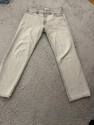 Ljusgrå raka jeans - Säljer ett par ljusgrå jeans med rak passform. Klassisk femficksmodell med knappgylf och bälteshällor. Jeansen har en enkel och clean look som passar till det mesta. Materialet är mjukt och bekvämt jeans-tyg.