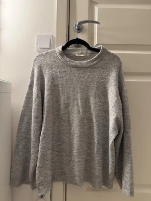 Ljusgrå oversized stickad tröja - Mysig ljusgrå stickad tröja med avslappnad passform och rund halsringning. Tröjan har långa ärmar och ribbade muddar vid ärmslut och nederkant. Perfekt för lager på lager och enkel att matcha med olika stilar. Passar för dig som brukar ha storlek S.