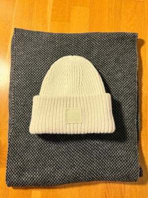 Acne Studios mössa - Säljer en beige/crémefärgad ribbstickad mössa från Acne Studios med uppvikt kant och det klassiska ansiktslogotypen framtill. Mössan är gjord i tjockt, mjukt material som håller dig varm samtidigt som den håller stilen och bekvämligheten ❄️🤩