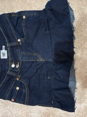 Mörkblå jeanskjol från 157 - Säljer en mörkblå jeanskjol från 157 med klassiska fickor fram och bak, samt rå kant nertill för en trendig look. Kjolen har kontrastsömmar i gult och knappgylf framtill. Perfekt för dig som gillar denim och vill ha en enkel men snygg kjol.