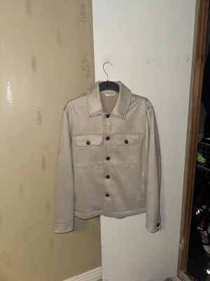 Beige Overshirt - Stilren beige jeansjacka i mockaimitation med klassisk krage och svarta knappar framtill. Två bröstfickor med lock och knapp samt raka sidfickor. Långärmad modell med enkel, clean look som passar till det mesta.