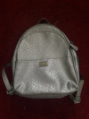 Silverfärgad ryggsäck från Guess (äkta)  - Snygg ryggsäck från Guess i silverfärgat syntetmaterial med GG-logga över hela väskan. Väskan har justerbara axelband, dragkedja upptill och en metallbricka med Guess-loggan på framsidan. Perfekt för dig som vill ha en trendig och praktisk accessoar.
