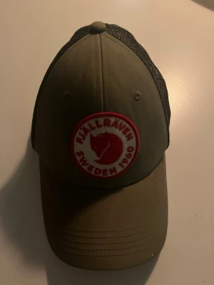 Olivgrön Fjällräven keps  - Snygg olivgrön keps från Fjällräven med klassisk röd och vit logga framtill. Kepsen har böjd skärm och svart mesh på sidorna och bak för extra ventilation. Justerbar baktill för perfekt passform. Kepsen är andvänd max 5 gånger, pris kan diskuteras vid snabb affär