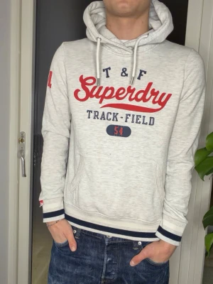 Ljusgrå hoodie från Superdry - Ljusgrå hoodie från Superdry med röd och marinblå text på bröstet och siffran 54 på ärmen. Klassisk känguruficka, dragsko i huvan och ribbade muddar med marinblå och vita ränder. Perfekt för en sportig och avslappnad stil. Jag på bilden är 177