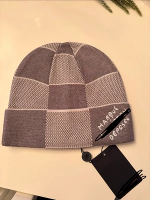 Grå-rutig  mössa  - Säljer en snygg grå och beige stickad mössa med rutigt mönster och broderad text framtill. Skriv om du vill ha mer bilder där det inte är ritat över. Helt oanvänd jätte fin skick 