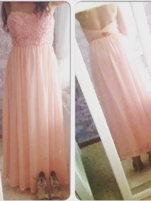 Ljusrosa långklänning med spetsdetaljer - Romantisk ljusrosa långklänning med axelbandslös design och vackert blommigt spetsliv. Kjolen är mjukt fallande i tunt material och klänningen har öppen rygg med rosettdetalj. Perfekt för speciella tillfällen. 