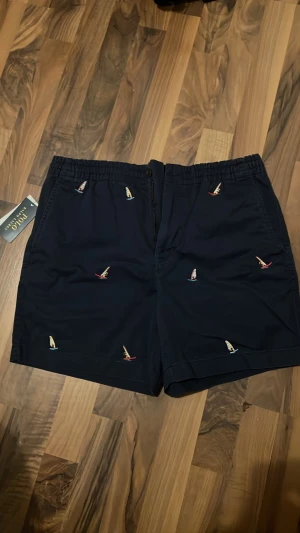 Marinblå shorts med broderade segelbåtar från Ralph Lauren - Oanvända marinblå shorts från Polo Ralph Lauren med broderade små segelbåtar i olika färger. Shortsen har elastisk midja, sidofickor och en bakficka med knapp. Perfekta för sommardagar och strandhäng. Original pris 1495kr