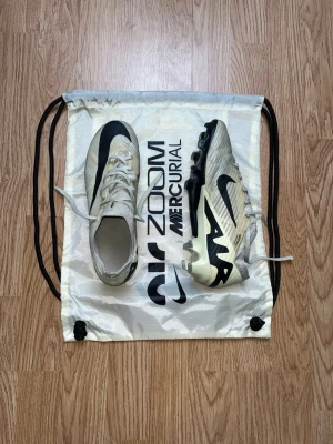 Nike Air Zoom Mercurial fotbollsskor - Säljer ett par Nike Air Zoom Mercurial fotbollsskor i vitt och svart med snygg, modern design. Skorna har snörning, platt sula med dobbar och är tillverkade i syntetmaterial för bästa prestanda på planen. Perfekta för dig som vill ha stil och funktion på matchen. Storlek 39. Bara hör av er vid funderingar!😃
