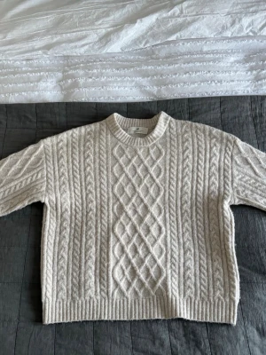 Beige stickad tröja från H&M med detaljer - Säljer en beige stickad tröja från H&M med klassiskt flätmönster och rund halsring. Tröjan har långa ärmar och ribbade muddar vid ärmslut och nederkant. Perfekt för kalla dagar och superenkel att styla till jeans eller kjol.